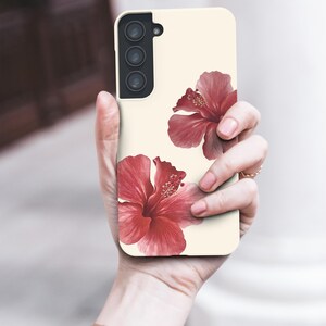May include: A phone case with a cream-colored background featuring two red hibiscus flowers. The case has a camera module with three lenses. The phone case is held in a person's hand, with a gold watch on the wrist.