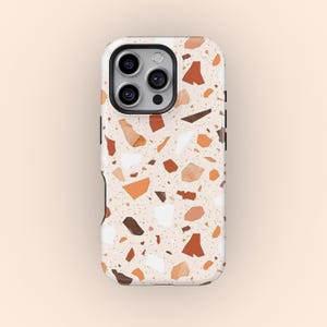 May include: A phone case with a terrazzo design. The case features a cream-colored background with various shapes in shades of brown, orange, and white. The case has a camera cutout and black side accents.