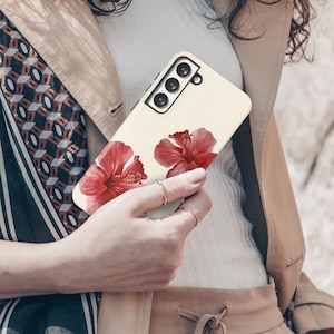 May include: A cream-colored phone case featuring two vibrant red hibiscus flowers. The case has a camera cutout and is held by a person wearing rings and a tan jacket.