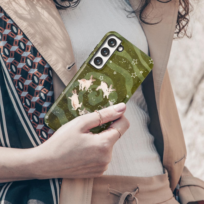Puede incluir: Una funda de tel&eacute;fono verde con un dise&ntilde;o caprichoso de conejos y estrellas. La funda tiene un patr&oacute;n de remolino y un recorte para la c&aacute;mara. La funda del tel&eacute;fono la sostiene una persona.