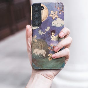 May include: A phone case featuring a whimsical illustration with a unicorn, moon, stars, and clouds against a purple background. The case is held in a hand, showcasing the design's details and colors.