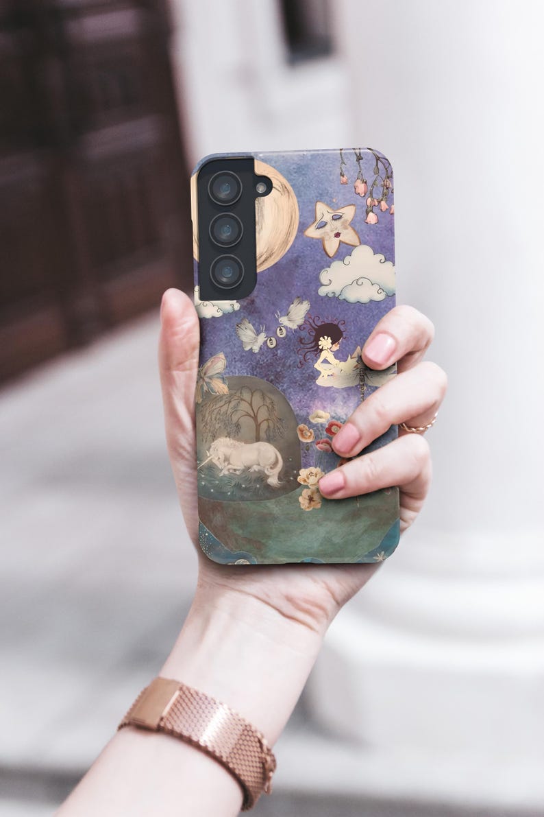May include: A phone case featuring a whimsical illustration with a unicorn, moon, stars, and clouds against a purple background. The case is held in a hand, showcasing the design's details and colors.