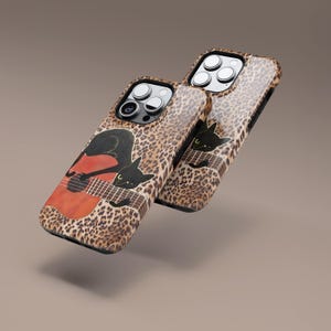 Może przedstawiać: Dwa etui na telefon z nadrukiem w cętki. Każde etui przedstawia czarnego kota grającego na czerwono-brązowej gitarze. Etui są ustawione pod kątem, prezentując wz&oacute;r i aparat telefonu.