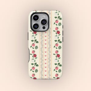 Puede incluir: Una funda de teléfono blanca con un patrón de fresas rojas y verdes, flores blancas y corazones rosas. La funda tiene un borde negro y un recorte para la cámara del teléfono.