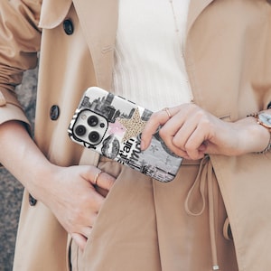 May include: A phone case with a black and white cityscape design, a pink star, and a leopard print star. The case also features the text "t'ain aime" and a pair of lips. The case is held by a person wearing a beige trench coat.