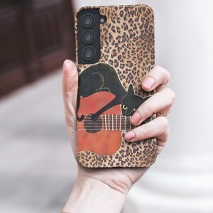 Może przedstawiać: Etui na telefon z nadrukiem w cętki. Etui przedstawia czarnego kota grającego na czerwonej gitarze. Etui na telefon jest trzymane w dłoni osoby.