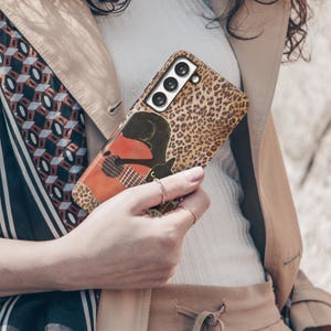 Może przedstawiać: Etui na telefon z nadrukiem w cętki lamparta i czarnym kotem grającym na gitarze. Etui ma brązowo-beżową kolorystykę. Etui na telefon jest trzymane przez osobę.