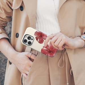 May include: A cream-colored phone case featuring two large red hibiscus flowers. The case has a glossy finish and is held in front of a tan trench coat.