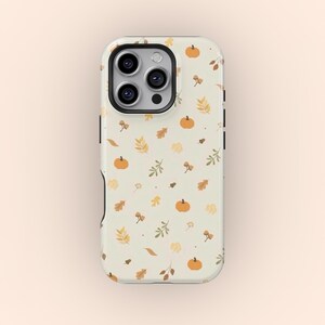 Puede incluir: Una funda de teléfono con un fondo crema con un patrón repetido de pequeñas calabazas naranjas, hojas de otoño y ramitas. La funda tiene un módulo de cámara negro y botones laterales negros.