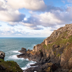 Puede incluir: Paisaje costero con un acantilado escarpado y estructuras de piedra históricas. El océano turquesa se encuentra con la costa rocosa bajo un cielo nublado. Vista panorámica de la costa de Cornualles.