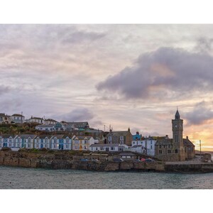 May include: A scenic view of a coastal town with a clock tower, houses, and a cloudy sky at sunset. The image shows a row of houses with blue and white facades along the coast, creating a picturesque waterfront scene.