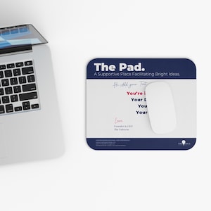 以下が含まれることがあります： ネイビーブルーの縁取りと「The Pad.」と「A Supportive Place Facilitating Bright Ideas.」の文字が入った白いマウスパッド。白いコンピューターマウスがパッドの上にあります。パッドには「You're i」、「Your L」、「You」、「Your」の文字も入っています。