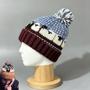 Könnte beinhalten: Eine Strickmütze mit dunkelweinrotem Rippbündchen und einem blauen Oberteil, das mit einem sich wiederholenden Muster aus weißen und schwarzen Schafen verziert ist. Die Mütze hat einen großen, runden, weiß-blauen Bommel aus dicken Garnfasern.