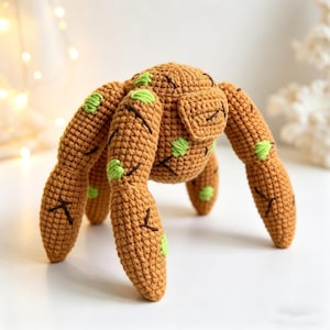 May include: A handmade crochet spider toy in shades of brown and green. The spider has eight legs, a rounded body, and green accents. The toy is made with a textured stitch and has embroidered details.
