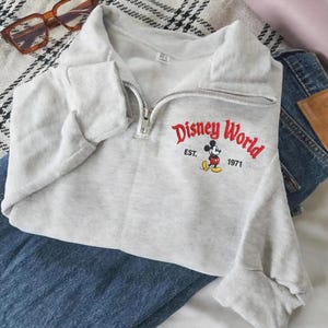 Puede incluir: Una sudadera gris claro con cuello alzado y cremallera. La parte delantera presenta el texto "Disney World" en rojo, con "EST. 1971" y un dibujo de Mickey Mouse. La sudadera est&aacute; doblada sobre unos vaqueros azules.