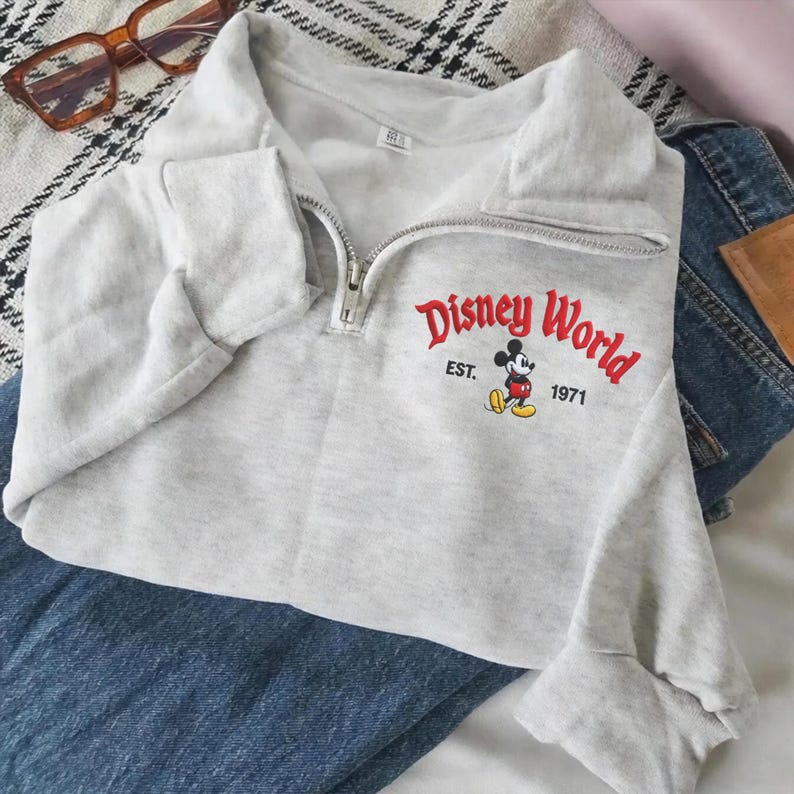 Puede incluir: Una sudadera gris claro con cuello alzado y cremallera. La parte delantera presenta el texto "Disney World" en rojo, con "EST. 1971" y un dibujo de Mickey Mouse. La sudadera est&aacute; doblada sobre unos vaqueros azules.