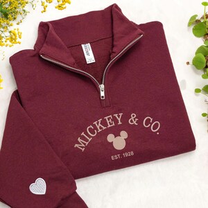 Puede incluir: Una sudadera burdeos con cremallera de un cuarto con el texto bordado "MICKEY & CO." y un dise&ntilde;o de cabeza de Mickey Mouse. La sudadera tambi&eacute;n presenta el texto "EST. 1928" y un peque&ntilde;o coraz&oacute;n en la manga.