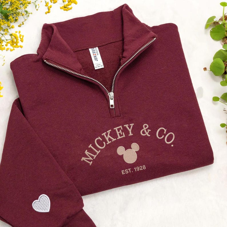 Puede incluir: Una sudadera burdeos con cremallera de un cuarto con el texto bordado "MICKEY & CO." y un dise&ntilde;o de cabeza de Mickey Mouse. La sudadera tambi&eacute;n presenta el texto "EST. 1928" y un peque&ntilde;o coraz&oacute;n en la manga.