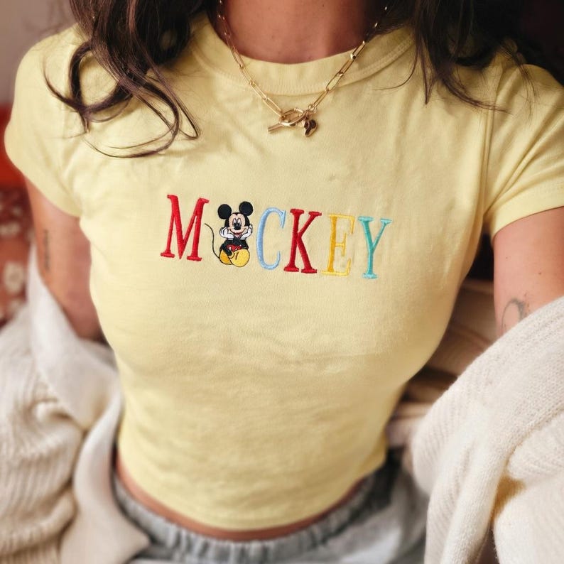 May include: A light yellow t-shirt with the embroidered word "Mickey" in red, black, yellow, and blue letters. The "o" in Mickey is replaced with a cartoon image of Mickey Mouse. The shirt has short sleeves and a round neckline.
