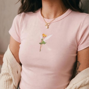 Puede incluir: Camiseta rosa claro con cuello redondo y bordado de hada con vestido verde y alas blancas. Se lleva un collar dorado con un colgante de cerradura. La camiseta tiene mangas cortas.