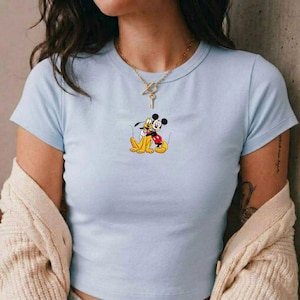 Puede incluir: Camiseta azul claro con un dibujo de Mickey Mouse abrazando a Pluto. Mickey lleva pantalones cortos rojos y Pluto es amarillo. La camiseta tiene cuello redondo y mangas cortas.