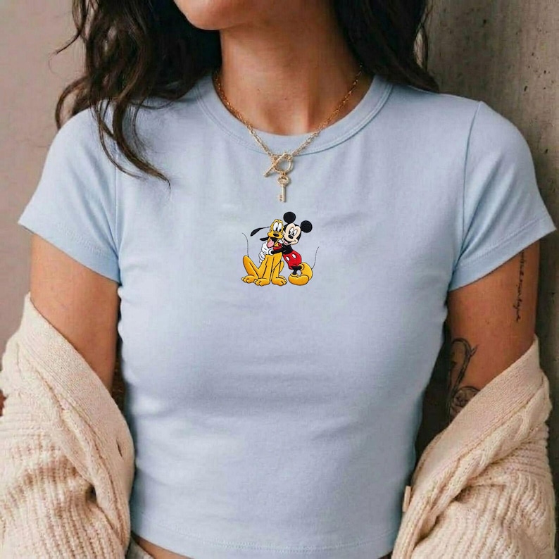 Puede incluir: Camiseta azul claro con un dibujo de Mickey Mouse abrazando a Pluto. Mickey lleva pantalones cortos rojos y Pluto es amarillo. La camiseta tiene cuello redondo y mangas cortas.