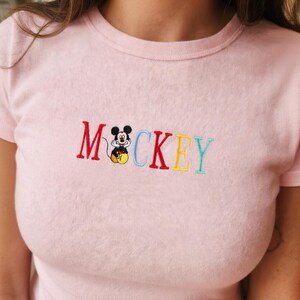 May include: A light pink t-shirt with the word "Mickey" embroidered in colorful letters. The letter "M" is red, "C" is light blue, "K" is red, "E" is yellow, and "Y" is teal. A cartoon Mickey Mouse is embroidered between the "M" and "C".