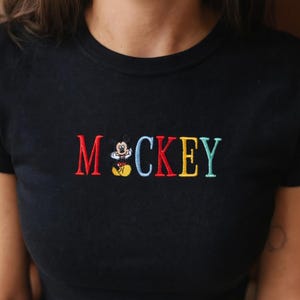 May include: Black crew neck t-shirt with embroidered "Mickey" in colorful letters. The "M" is red, the "C" is light blue, the "K" is red, the "E" is yellow, and the "Y" is teal. A small embroidered Mickey Mouse is between the "M" and "C".