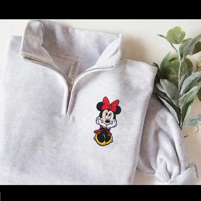May include: A light gray pullover sweatshirt with a quarter-zip neckline. The sweatshirt features an embroidered image of Minnie Mouse wearing a red bow and dress. The sleeves are rolled up, and a sprig of greenery is visible in the background.
