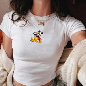 Puede incluir: Camiseta blanca con un gr&aacute;fico de Mickey Mouse y Pluto abraz&aacute;ndose. La camiseta tiene mangas cortas y cuello redondo. Se lleva un collar de cadena dorada. La camiseta est&aacute; hecha de un material suave.