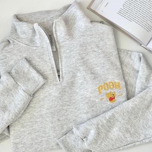 May include: A light grey quarter-zip pullover sweatshirt. The sweatshirt has the embroidered word "POOH" in gold, with a Winnie the Pooh graphic. The sleeves are long with ribbed cuffs. The garment is made of a soft material.