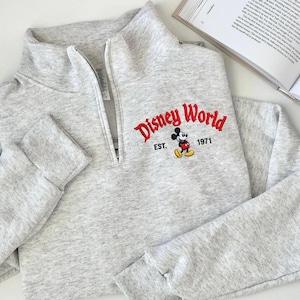 Puede incluir: Una sudadera gris claro con cuello con cremallera de un cuarto. La parte delantera presenta el texto bordado "Disney World" en rojo, con "EST. 1971" y un gr&aacute;fico de Mickey Mouse. La sudadera tiene mangas largas.