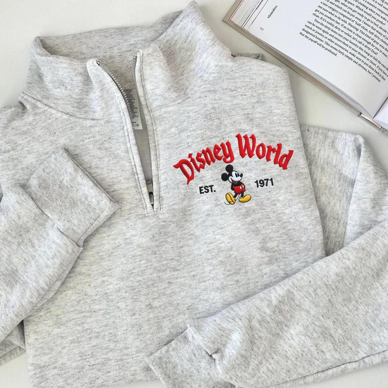 Puede incluir: Una sudadera gris claro con cuello con cremallera de un cuarto. La parte delantera presenta el texto bordado "Disney World" en rojo, con "EST. 1971" y un gr&aacute;fico de Mickey Mouse. La sudadera tiene mangas largas.
