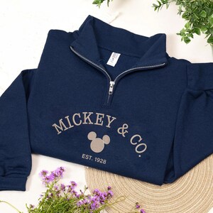 Puede incluir: Sudadera azul marino con cuello con cremallera. El texto bordado "MICKEY & CO." est&aacute; sobre un gr&aacute;fico de la cabeza de Mickey Mouse, con "EST. 1928" debajo. La sudadera est&aacute; sobre una superficie clara con flores moradas y vegetaci&oacute;n.