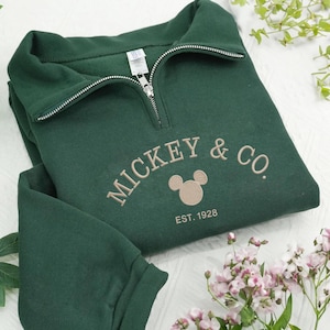 Puede incluir: Sudadera verde oscuro con cuello con cremallera de un cuarto. La sudadera presenta el texto bordado "MICKEY & CO." con un gráfico de la cabeza de Mickey Mouse y "EST. 1928". La sudadera está doblada y tiene un aspecto suave.