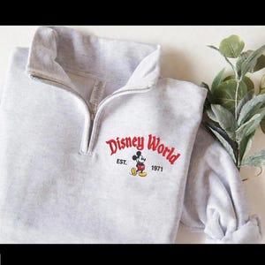 Puede incluir: Sudadera gris claro con cremallera de un cuarto de cuello, con las palabras "Disney World" en letras rojas, Mickey Mouse y el texto "EST. 1971". La sudadera tiene cuello alto y cremallera plateada.