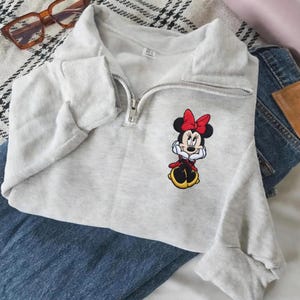 May include: A light gray sweatshirt with a quarter-zip neckline and a Minnie Mouse embroidered patch. The patch features Minnie in a red bow, dress, and yellow shoes. The sweatshirt is paired with blue jeans.