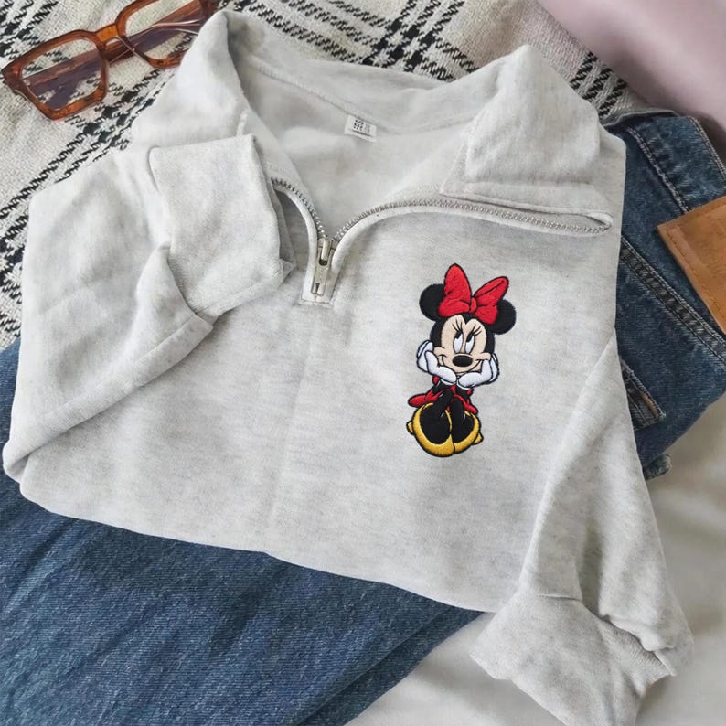 May include: A light gray sweatshirt with a quarter-zip neckline and a Minnie Mouse embroidered patch. The patch features Minnie in a red bow, dress, and yellow shoes. The sweatshirt is paired with blue jeans.