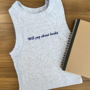 Puede incluir: Una camiseta sin mangas gris claro con el texto bordado "Will yap about books" en azul marino. Un cuaderno de espiral con una cubierta marrón está al lado de la camiseta sin mangas. La camiseta sin mangas está sobre una superficie de madera.