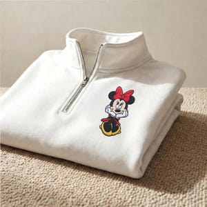 May include: White quarter-zip sweatshirt featuring a Minnie Mouse embroidered graphic. The sweatshirt has a high collar and a silver zipper. The Minnie Mouse graphic includes a red bow, black ears, and a red and black outfit.