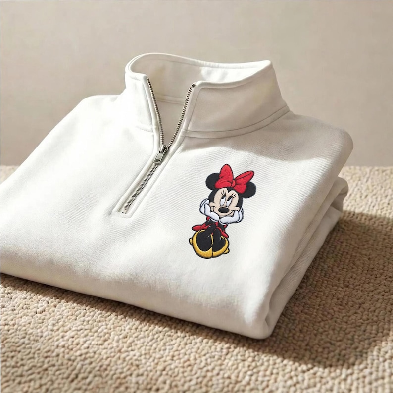 May include: White quarter-zip sweatshirt featuring a Minnie Mouse embroidered graphic. The sweatshirt has a high collar and a silver zipper. The Minnie Mouse graphic includes a red bow, black ears, and a red and black outfit.