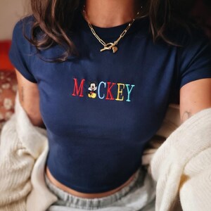 May include: Navy blue crop top with the embroidered word "Mickey" in red, yellow, blue, and green. The letter "M" is red, and the letter "C" is yellow. The top has a small Mickey Mouse graphic.