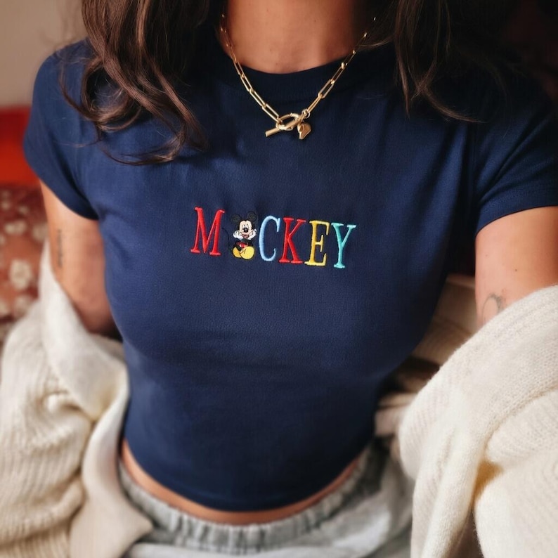 May include: Navy blue crop top with the embroidered word "Mickey" in red, yellow, blue, and green. The letter "M" is red, and the letter "C" is yellow. The top has a small Mickey Mouse graphic.