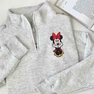 May include: A light gray sweatshirt with a quarter-zip neckline and long sleeves. The sweatshirt features an embroidered Minnie Mouse design on the front, with a red bow, red dress, and yellow shoes. The garment is made of a soft, comfortable material.