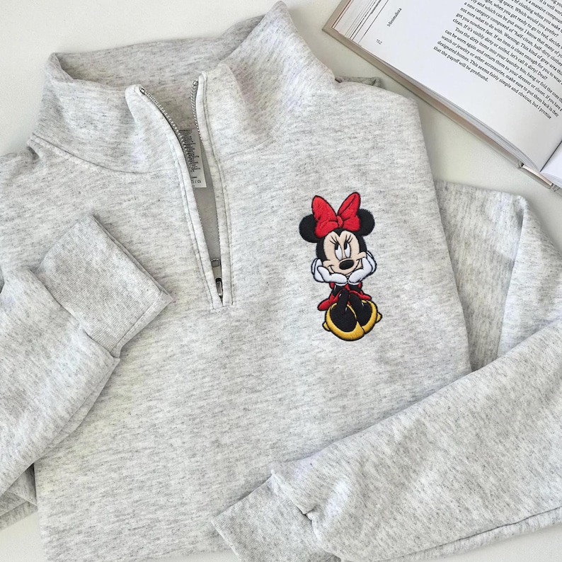 May include: A light gray sweatshirt with a quarter-zip neckline and long sleeves. The sweatshirt features an embroidered Minnie Mouse design on the front, with a red bow, red dress, and yellow shoes. The garment is made of a soft, comfortable material.