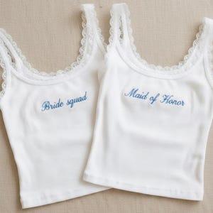 May include: Two white vest tops with lace trim. One has "Bride squad" embroidered in blue, the other says "Maid of Honor". The tops are made of a ribbed fabric.