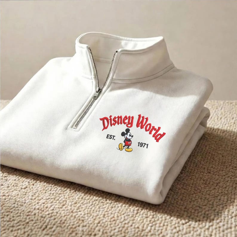 Puede incluir: Sudadera blanca con cuello con cremallera. La parte delantera presenta las palabras "Disney World" en rojo, con "EST. 1971" debajo, y una imagen de Mickey Mouse. La sudadera est&aacute; doblada sobre una superficie texturizada.