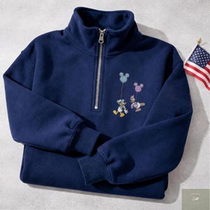Puede incluir: Sudadera azul marino con cremallera de cuarto de cremallera y personajes de Disney bordados. El diseño presenta a Pato Donald y Daisy sosteniendo globos con forma de Mickey Mouse. Una bandera americana está en el fondo.