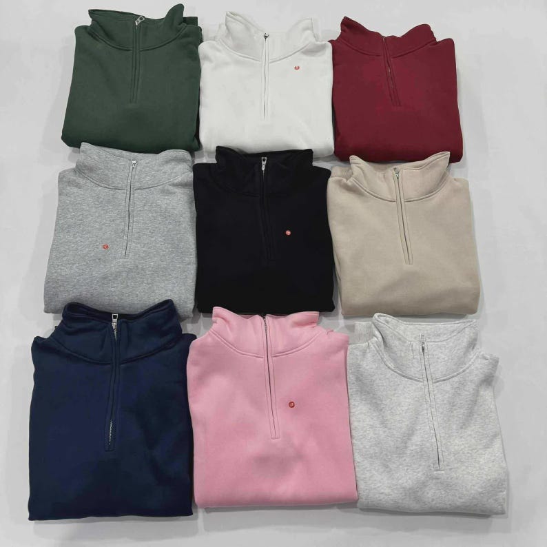 Puede incluir: Nueve sudaderas plegadas con cremallera de cuarto de cierre en varios colores: verde oscuro, blanco, burdeos, gris, negro, beige, azul marino, rosa y gris claro. Cada sudadera tiene un peque&ntilde;o logotipo rojo cerca de la cremallera.
