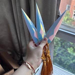 May include: Three iridescent kunai knives with gold tassels are held in a hand. The knives have a sharp, pointed design and reflect a range of colours. The tassels are a warm, golden-orange colour.